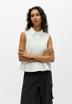 Femme aux cheveux bouclés et foncés portant une chemise blanche sans manches boutonnée et un pantalon noir à jambes larges, debout les bras croisés devant un fond blanc.
