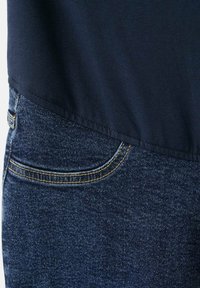 Donkerblauwe stof die overlapt op blauwe spijkerbroek met zichtbare gestikte zak en riemlus.