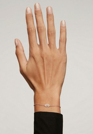 Mano con uñas naturales que lleva una delicada pulsera de plata con un charm ovalado, sobre un fondo claro y liso.