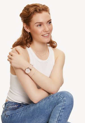 Rose gold watch with a round face and light pink strap, worn with layered silver necklaces on a model in a white tank top and jeans.