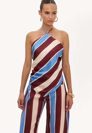 Femme portant un top sans manches à col licou et un pantalon large à rayures diagonales et verticales bordeaux, bleu et crème, accessoirisée de boucles d’oreilles créoles dorées.