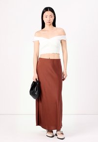 Hvit off-the-shoulder crop topp med volanger, kombinert med et langt brunt satengskjørt og svart håndveske. Fottøyet har lyse stropper.