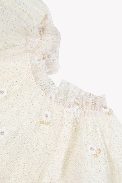 Ύφασμα μουσελίνας σε κρεμ απόχρωση με χρυσές νιφάδες, που διαθέτει λαιμόκοψη με βολάν και διακοσμητικά με κεντήματα λουλουδιών. Μαλακό και υφασμένο, με διαφανή ποιότητα.