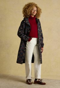 Garšs, rakstains mētelis tumši zilā krāsā ar ziedu rakstu, sarkans augsta kakla džemperis, balti bikses un brūni apavi ar akcentiem.