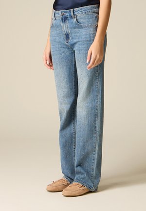 Mujer vestida con vaqueros rectos azul claro con pequeños remaches metálicos, combinados con mocasines color marrón claro sobre un fondo beige liso.