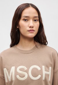 Καφέ φούτερ με μεγάλα, κρεμ χρώματος κεντημένα γράμματα "MSCH," με στρογγυλή λαιμόκοψη και μαλακή υφή υφάσ ματος.