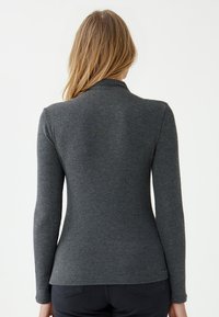 Vrouw met lang blond haar, gekleed in een strakke donkergrijze langslavend top en zwarte broek, gezien van achteren tegen een witte achtergrond.