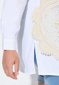Witte blouse met een gehaakt beige kantdetail aan de zijkant. Voorzien van een manchet met knopen en een zijsplit. Zachte textuur en lichtgewicht stof.