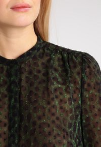 Blouse verte transparente à motifs avec col arrondi, pois texturés et fermeture par boutons, avec de subtils plis aux épaules.