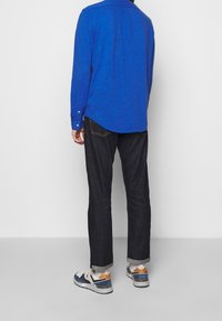 Blå långärmad skjorta med knappdetaljer, kombinerad med mörka denimjeans och vita sneakers med navy- och orangeaccenter.