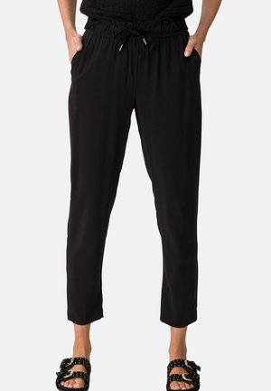 Femme portant un pantalon noir à taille élastique avec cordon de serrage et des sandales noires à enfiler avec boucles dorées, debout les mains dans les poches.