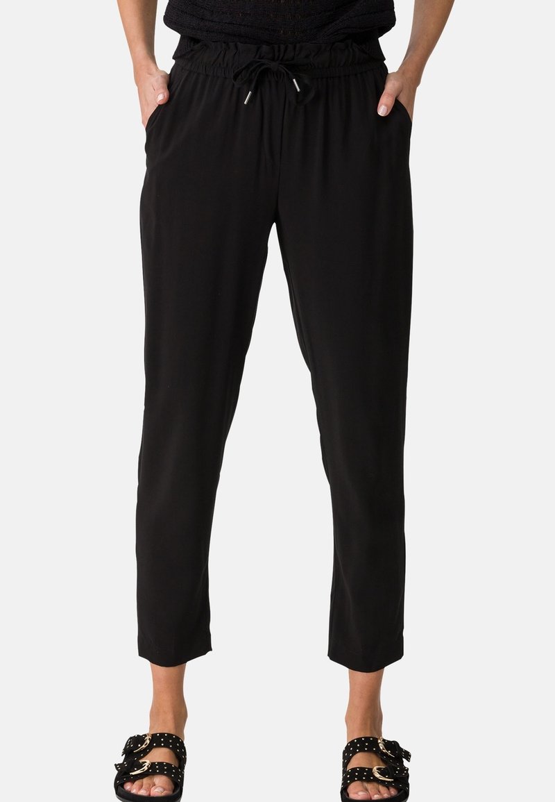 Femme portant un pantalon noir à taille élastique avec cordon de serrage et des sandales noires à enfiler avec boucles dorées, debout les mains dans les poches.