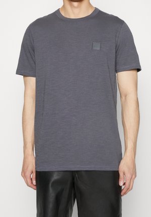 Mężczyzna w gładkim ciemnoszarym T-shircie z krótkim rękawem z małym kwadratowym logo na lewej piersi, w połączeniu z czarnymi spodniami.