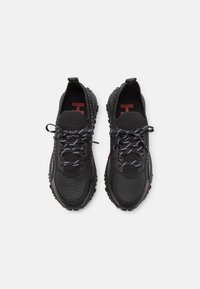 Černé sportovní boty s texturovaným síťovaným svrškem, kontrastními bílými a černými tkaničkami a robustními gumovými podrážkami. Červené akcenty loga.