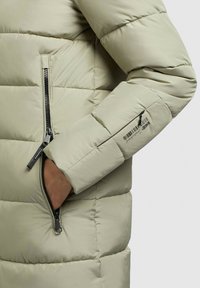 Chaqueta de pluma verde claro con textura acolchada, bolsillos con cremallera y detalles en metal negro. La manga incluye un detalle de la marca.