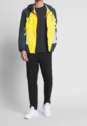 Veste coupe-vent jaune et canard, avec une capuche et des poignets élastiques. Associée à un pantalon noir et des chaussures de sport blanches.
