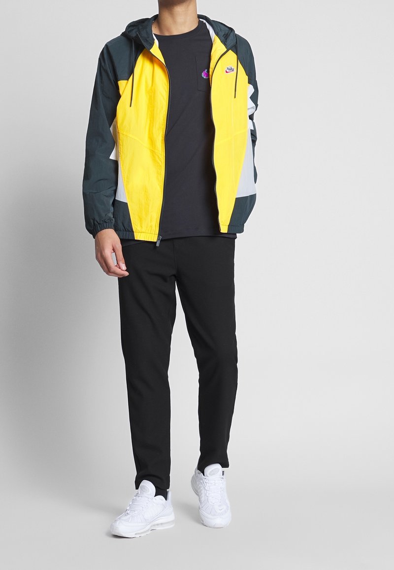 Windbreaker jacket in yellow and teal, featuring a hood and elastic cuffs. Paired with black pants and white athletic shoes.