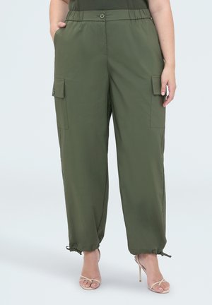 Mujer con pantalones cargo verde oliva con tobillos ajustables mediante cordón y sandalias de tacón alto beige, de pie con las manos en los bolsillos.