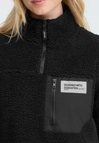 Zwarte fleece pullover met een hoge kraag, ritssluiting aan de voorkant, en een ritszak op de borst met een label en logo. Textuur, warm materiaal.