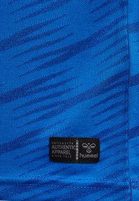 Tissu bleu avec un motif ondulé texturé, portant une étiquette noire marquée "Hummel" et "Authentic Apparel" en blanc.