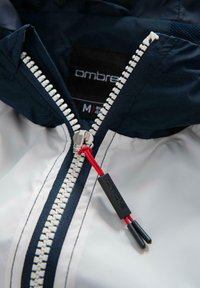 Hvid og marineblå zip-jacket med marineblå krave, der har en fremtrædende hvid lynlås og rød detalje på lynlås trækkeren. Mærkelabel synlig.