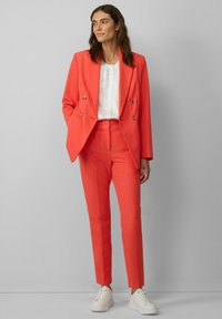 Korallenfarbener, doppelt zugeknöpfter Blazer kombiniert mit passenden, engen Hosen. Das Outfit wird durch eine weiße Bluse und weiße Sneakers abgerundet.