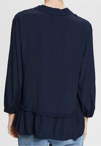 Blouse bleu marine en tissu léger, dotée d'un ourlet à volants, de manches longues avec détails boutonnés et d'un col classique.