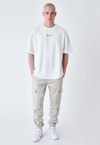 Un giovane con i capelli biondi corti che indossa una maglietta bianca oversize, pantaloni cargo beige chiaro e sneakers bianche, in piedi su uno sfondo neutro.