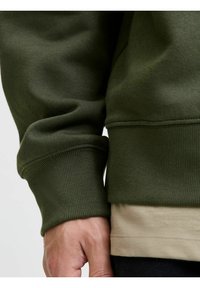 Olijfgroene sweatshirt met ribbelmanchetten en een gestructureerde stof. Gelaagd met een lichtbeige shirt eronder. Close-up van de mouw en hand.