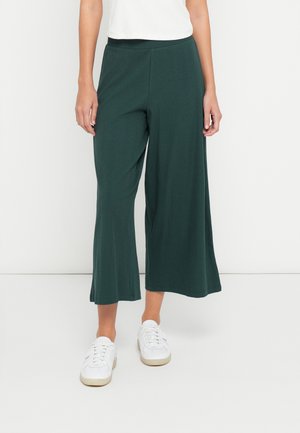 Person trägt dunkelgrüne, weit geschnittene kurze Hose mit weißem Oberteil und weißen Sneakers vor einem schlichten Hintergrund.