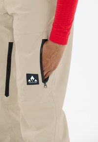 Beige wasserdichte Hose mit zwei seitlichen Reißverschlüssen und einem schwarzen Logo-Patch. Das Kleidungsstück zeichnet sich durch eine glatte, elegante Textur und verstärkte Nähte aus.