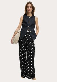 Denim-Trägershirt mit goldenen Knöpfen, kombiniert mit schwarzen weit geschnittenen Hosen mit weißen Punkten. Das Model hält eine beigefarbene gefaltete Tasche.