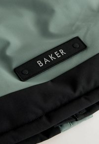 Zielono-czarna kurtka z prostokątną czarną naszywką z napisem "BAKER" w srebrnym kolorze. Gładka, matowa faktura z warstwowym wzornictwem.