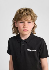 Svart pikétröja av texturerat tyg med krage, tre knappar och en vit logotyp på vänster bröst.