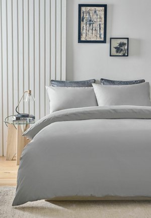 Modernes Schlafzimmer mit grauer Bettwäsche, zwei gerahmten abstrakten Gemälden an weißer Wand, Glastisch mit Lampe und Holzboden.