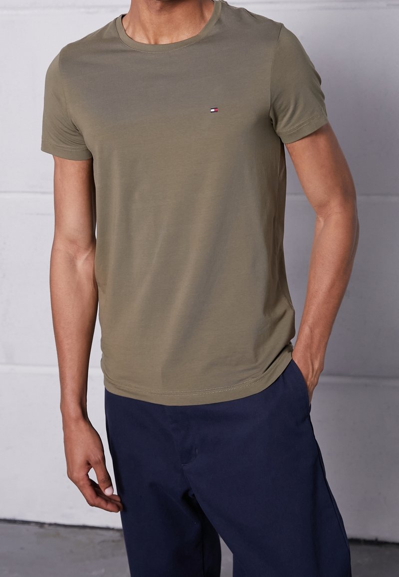 T-shirt vert olive à manches courtes en tissu doux, doté d'un col rond et d'un petit logo sur la poitrine, accompagné d'un pantalon bleu foncé.