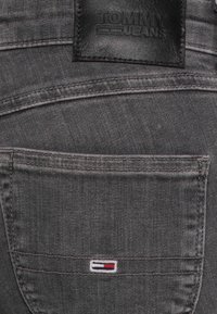Jeans de mezclilla gris con un bolsillo trasero que presenta un pequeño logotipo bordado de Tommy Jeans en rojo, blanco y azul, y un parche de logo de cuero negro.