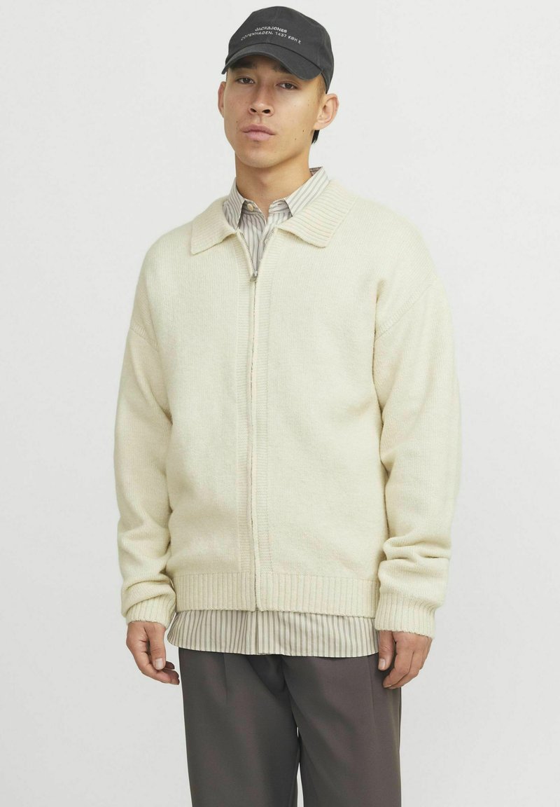 Cremfarbener gestrickter Cardigan mit einem Reißverschluss, gerippten Bündchen und Saum, über einem gestreiften Hemd getragen, kombiniert mit grauen Hosen und einer schwarzen Kappe.