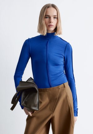 Langärmeliges Rollkragenoberteil in lebhaftem Blau, mit schlankem Design und dezenten Nahtdetails, kombiniert mit hoch taillierter brauner Hose und einer dunklen Tasche.