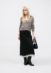 Cardigan gris clair à fermeture éclair porté sur une robe maxi noire avec un bord en dentelle à l'ourlet, accompagné de bottes noires montant jusqu'aux genoux et d'un sac à main noir.