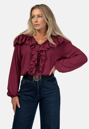 Bordeauxfarbene Bluse mit gerafftem Ausschnitt und Vorderseite, langen Ärmeln, aus leichtem Material. Kombiniert mit dunkelblauen Denim-Hosen und einem schwarzen Gürtel.