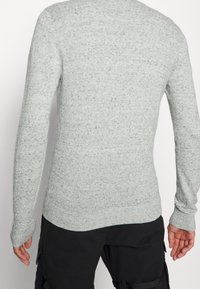 Světlý šedý svetr s dlouhými rukávy, texturovaným povrchem a žebrovanými manžetami. Má kulatý výstřih a přiléhavý střih.