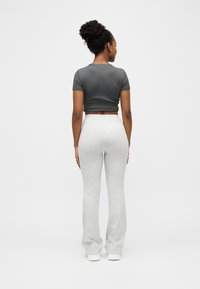 Femme aux cheveux bouclés attachés en chignon, portant un haut court gris foncé ajusté, un pantalon évasé gris clair et des baskets blanches, debout de dos sur fond blanc.