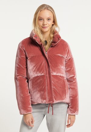 Jonge vrouw met lang blond haar die een roze fluwelen donsjasje draagt en lichtgrijze broek, staand voor een effen achtergrond.