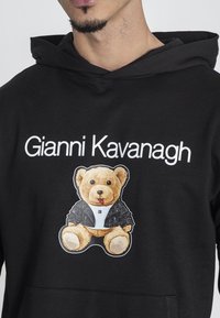 Sudadera negra con un gráfico de un oso que lleva chaqueta de cuero y camisa blanca. Texto "Gianni Kavanagh" mostrado de manera prominente sobre el gráfico.