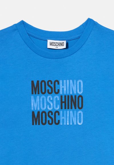 Μπλε παιδικό μπλουζάκι με το κείμενο "MOSCHINO" τυπωμένο τρεις φορές εναλλάξ με μαύρα και ανοιχτό μπλε γράμματα στο μπροστινό μέρος.