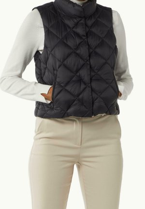 Personne portant un gilet matelassé noir sur un haut blanc à manches longues et un pantalon beige, les mains dans les poches du gilet.