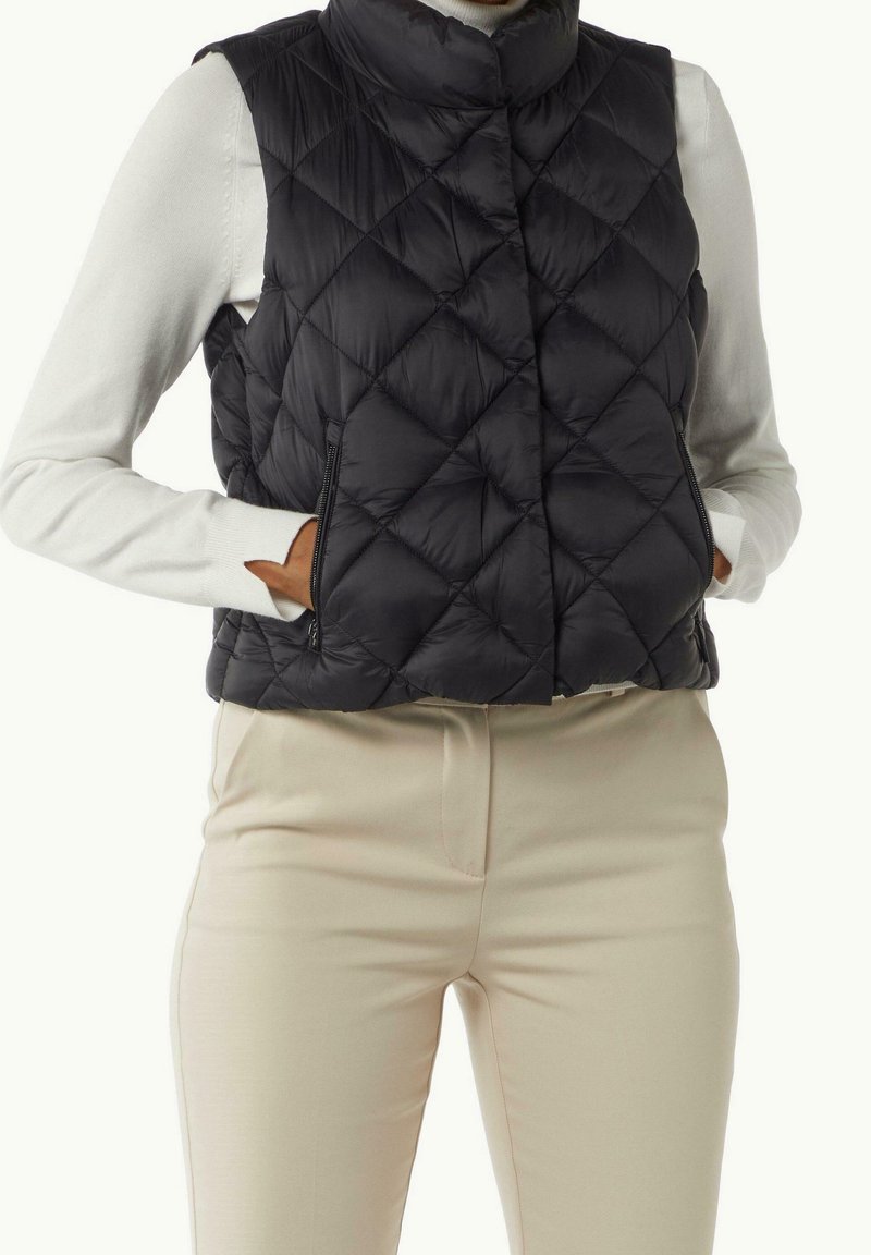 Personne portant un gilet matelassé noir sur un haut blanc à manches longues et un pantalon beige, les mains dans les poches du gilet.