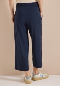 Personne portant un pantalon court bleu marine avec une ceinture élastique et des baskets blanches avec des accents bleus, debout devant un fond beige.