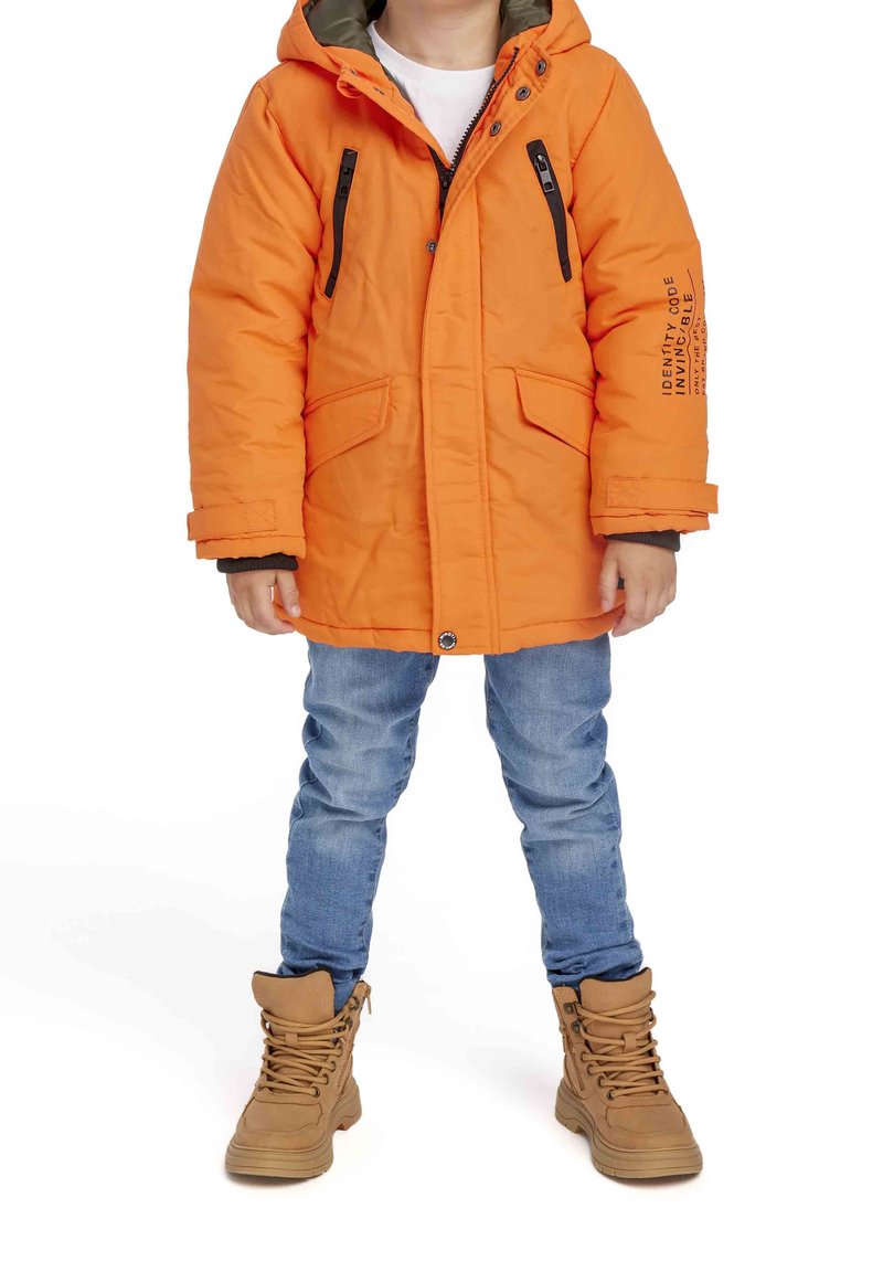 Manteau rembourré orange avec capuche, zippé noir et accents de poches, affichant un texte imprimé sur la manche. Associé à un jeans bleu et des bottes marron.